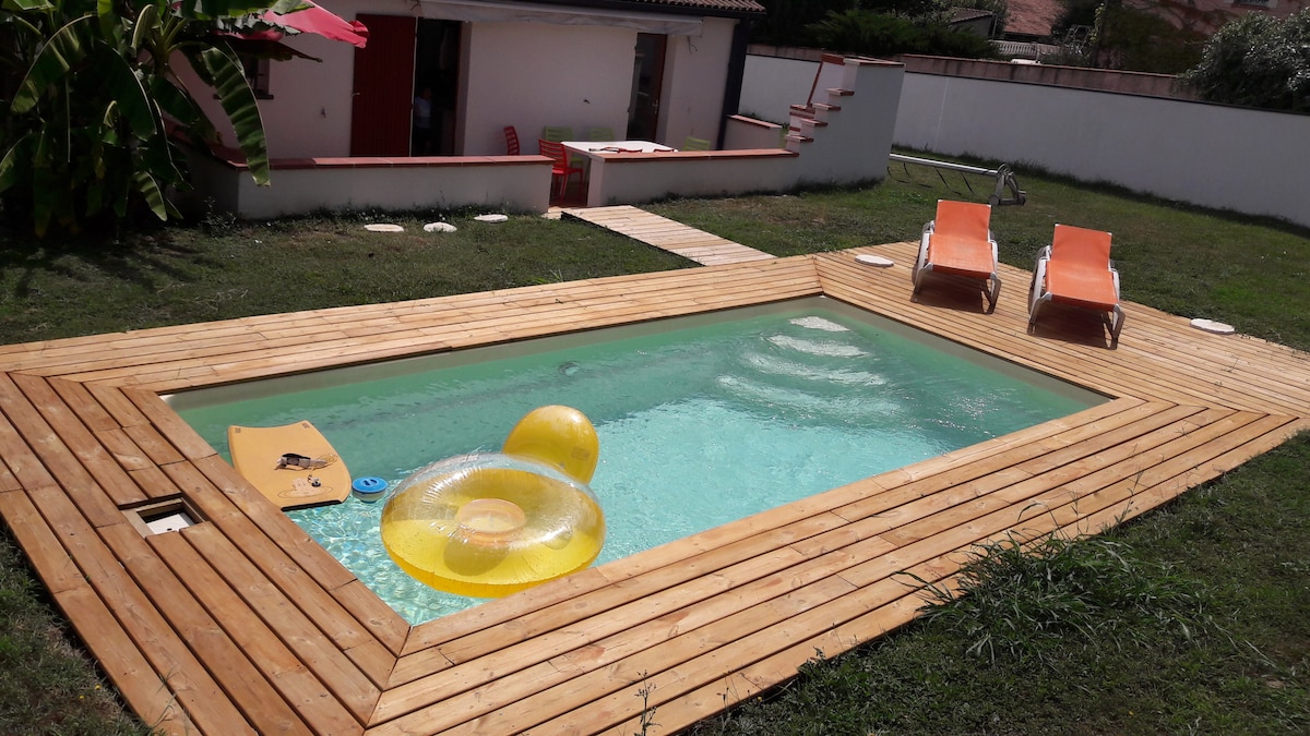 A modest swimming pool is surrounded by a wooden deck, which features two bright orange lounge chairs. A yellow floatation device rests in the water, while lush greenery borders the space, enhancing the outdoor setting.
