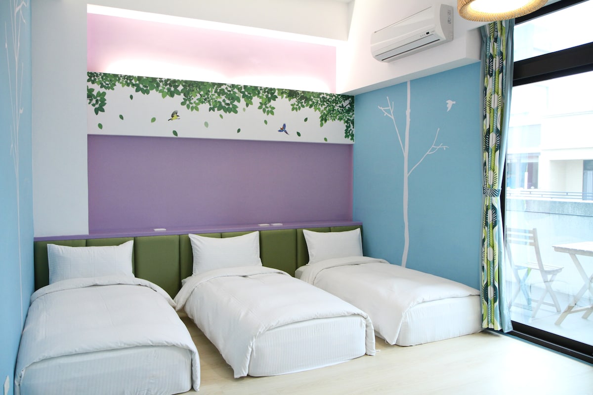 A spacious room is designed with three cozy beds arranged neatly across the floor. Light-colored walls feature soft, natural motifs, while large windows provide ample daylight, framed by vibrant curtains. The soothing color palette enhances a relaxed atmosphere.