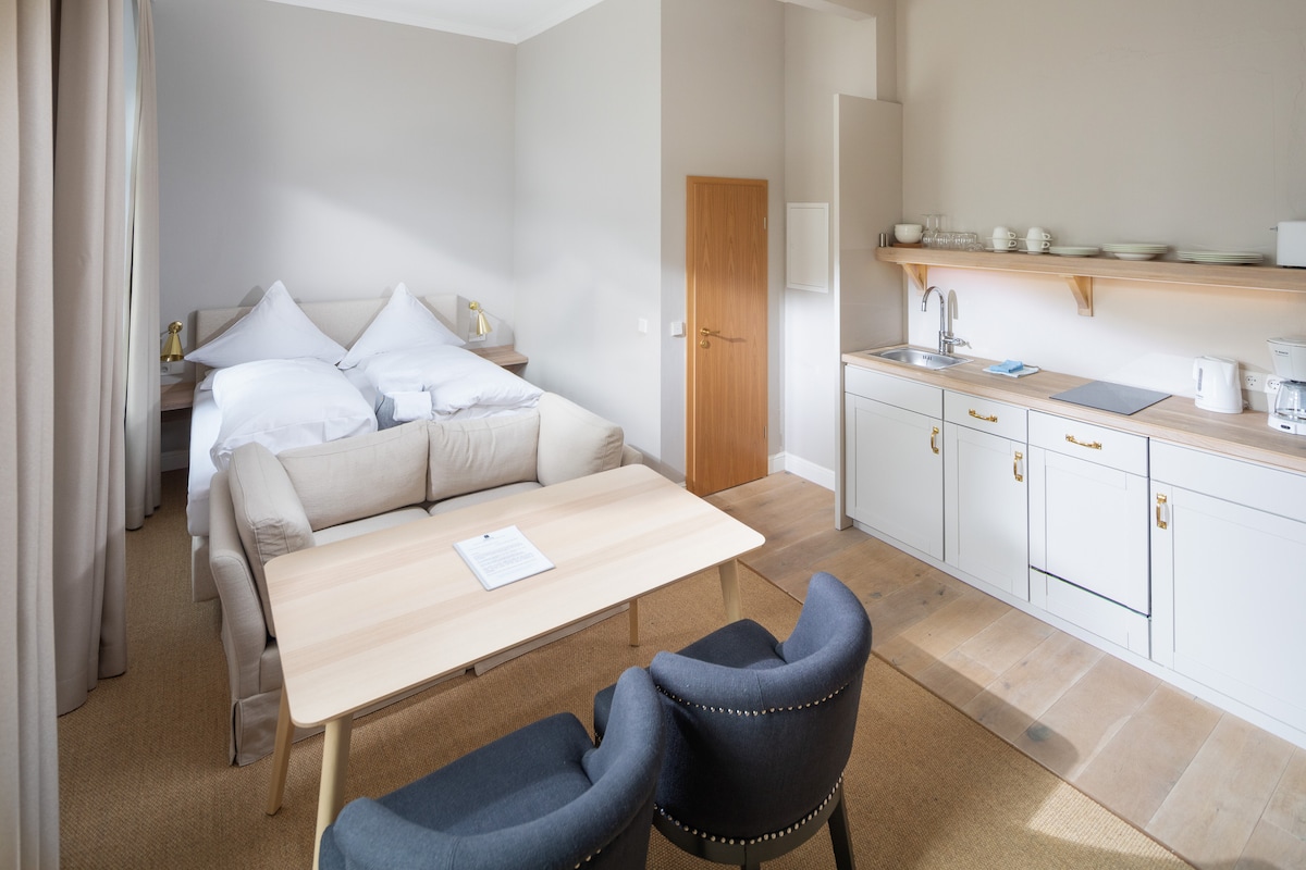 A spacious studio features a light and airy ambience with a cozy seating area, a contemporary table, and a well-equipped kitchenette. Two beds are neatly arranged against the wall, while natural light filters through large windows, creating a welcoming atmosphere.