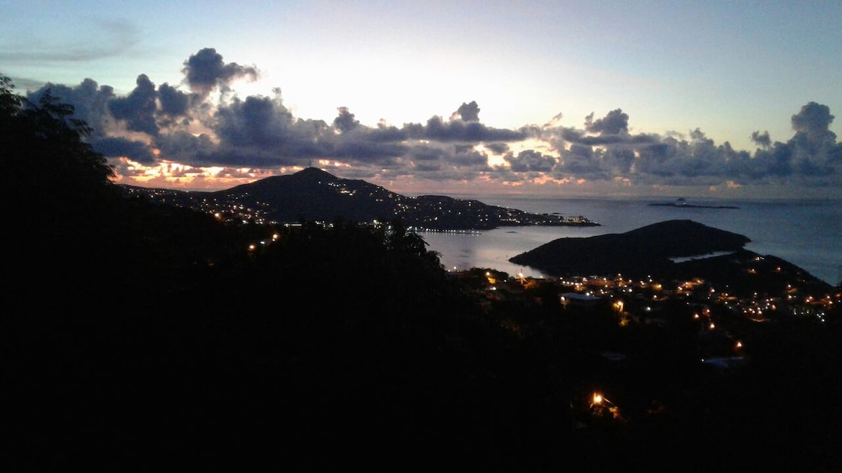 A scenic view captures the setting sun over Charlotte Amalie harbor, with silhouettes of surrounding hills and twinkling lights reflecting in the water. The sky transitions from orange to deep blue, framed by clouds, creating a serene atmosphere ideal for relaxation.