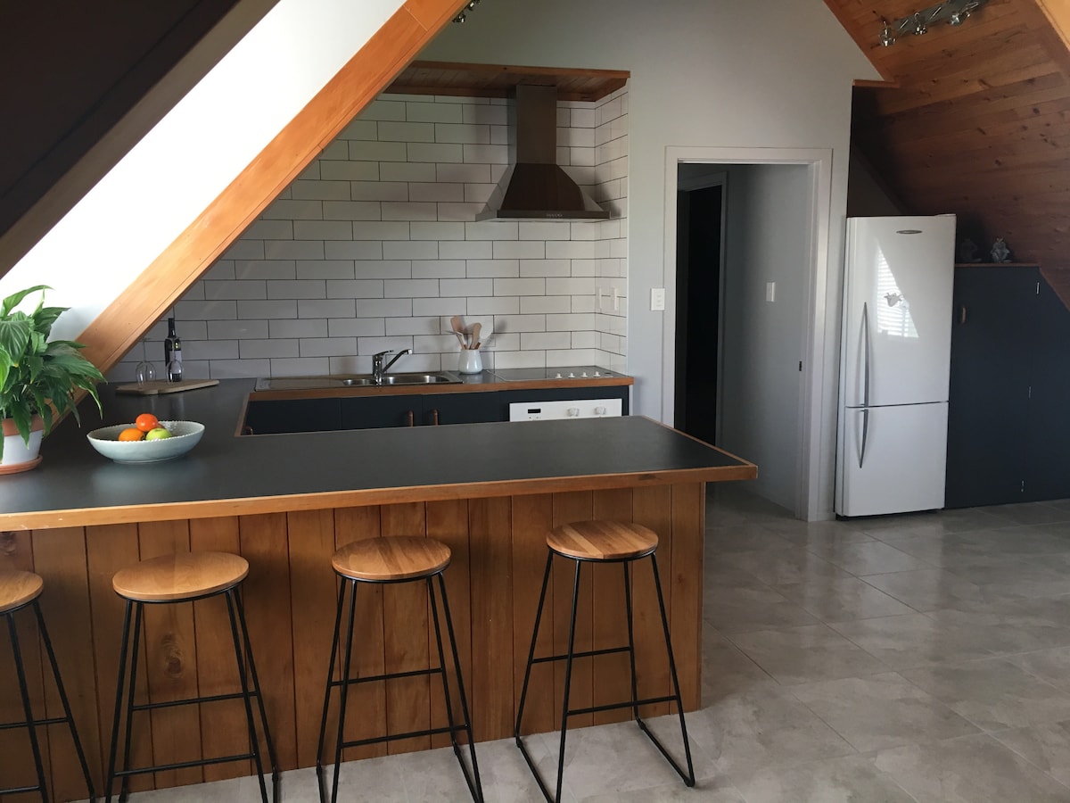 The kitchen features a modern design with wooden accents and a spacious island bar with three stools. A grey countertop contrasts with white tiles, and stainless steel appliances, including a refrigerator, are visible. A bowl of fresh fruit adds a touch of color on the island.