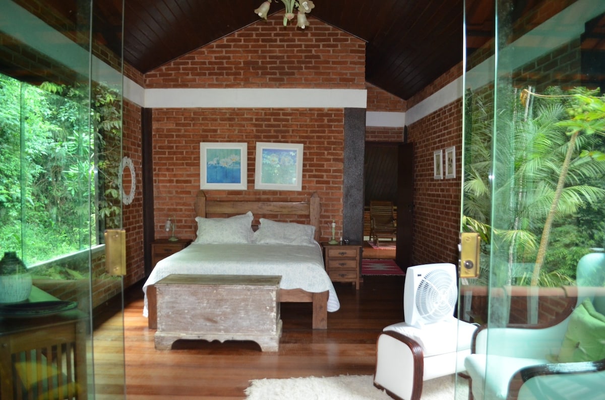 A spacious bedroom features exposed brick walls and large glass windows, providing views of the surrounding greenery. A queen bed is complemented by wooden furniture including nightstands and a rustic dresser. A chair and fan are also included, enhancing comfort in the serene atmosphere.