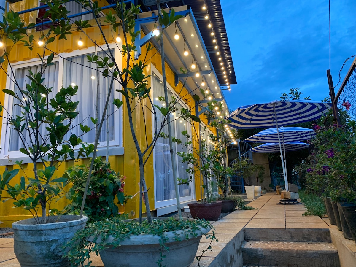 Soft lighting is displayed overhead, illuminating the yellow exterior of the house. Potted plants are arranged along a pathway, with striped umbrellas providing shade nearby. The scene is set in the early evening, showcasing the tranquil ambiance of the outdoor area.