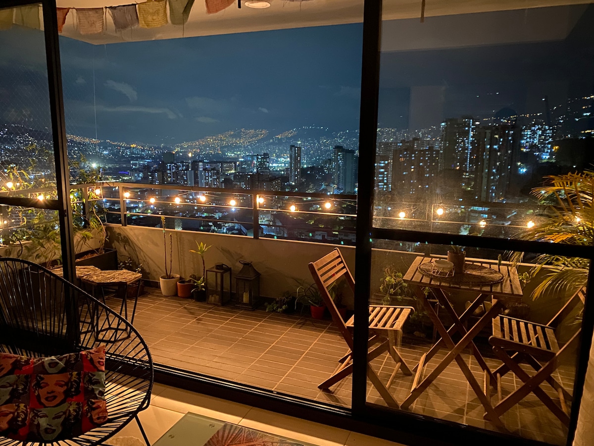 A balcony with a stunning city view is showcased at night, illuminated by soft string lights. Two wooden folding chairs and a small table are present, surrounded by potted plants. In the background, the lights of the city create a vibrant skyline.