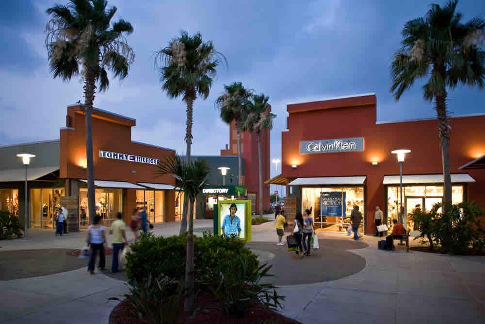 An outdoor shopping area is presented with storefronts from various brands, some featuring illuminated signs. Palm trees frame the scene, and a directory stand is located in front. The surroundings suggest an engaging shopping environment during twilight.