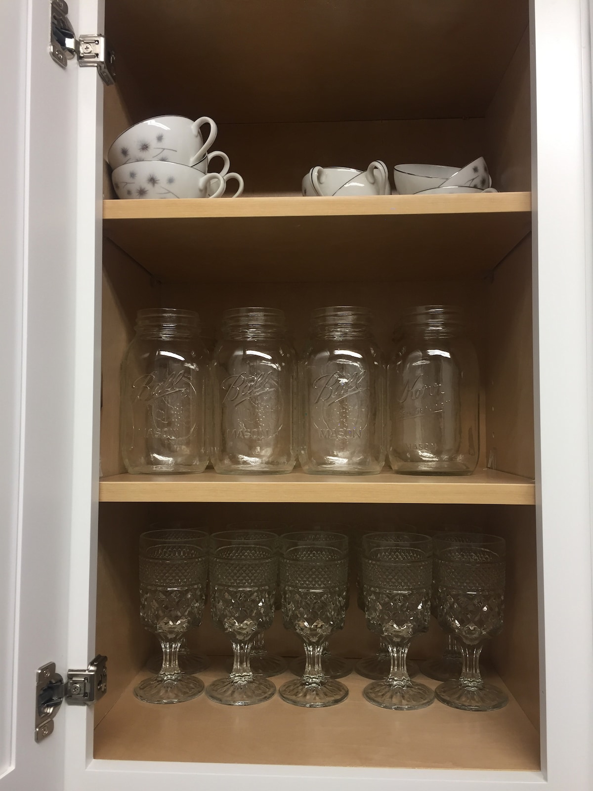 A neatly organized kitchen cabinet displays glassware and dining essentials. The top shelf holds delicate teacups and saucers, while the middle shelf contains mason jars. The bottom shelf showcases elegant glass goblets, highlighting a functional and stylish kitchen setup.
