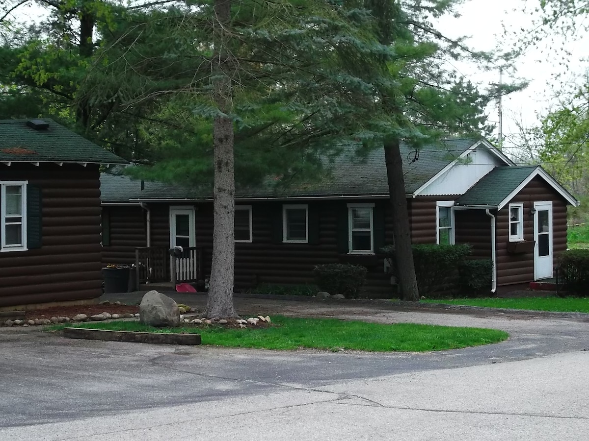 The exterior of a rustic, log cabin-style property is visible, set amidst lush greenery. The cabin features multiple entrances and includes large windows that invite natural light. A well-maintained lawn and decorative rocks add to the inviting outdoors.