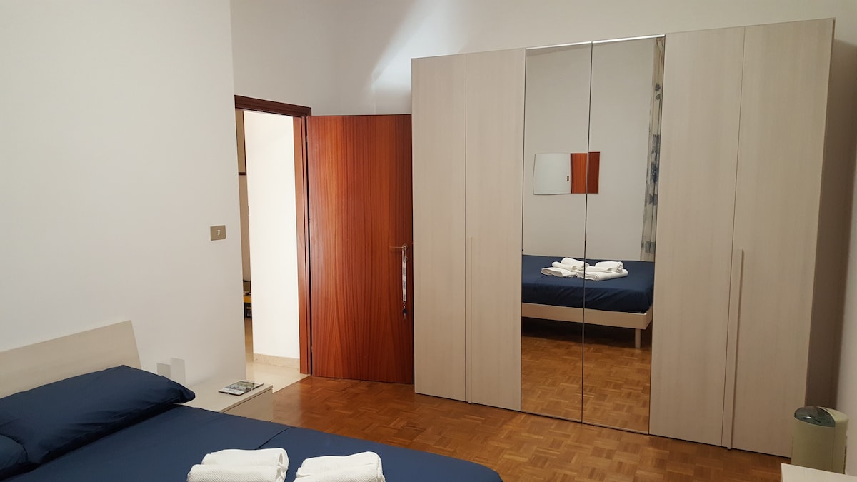 The room features a spacious bed with navy blue linens. A mirrored wardrobe reflects the surroundings, and a light-colored wood floor adds warmth. A doorway suggests entry to an adjoining space, while neatly arranged towels are placed on the bed.