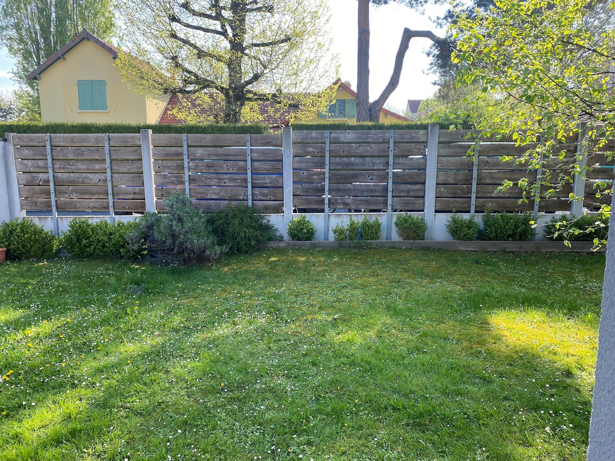 A well-maintained garden area is visible, featuring lush green grass and small hedges along the perimeter. A wooden fence provides a sense of privacy, with patches of sunlight filtering through the trees, creating a peaceful outdoor space.