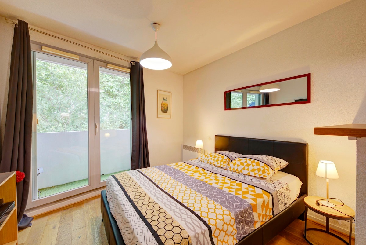 A well-designed bedroom features a spacious bed with patterned bedding, complemented by soft bedside lamps. Large glass doors provide access to a balcony overlooking greenery, enhancing natural light in the room. Decorative elements include a mirror and wall art, enriching the space.
