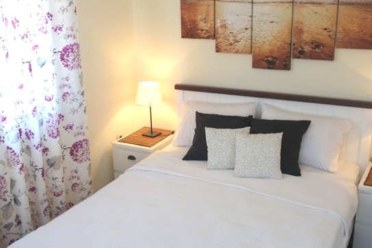A cozy bedroom features a spacious bed with crisp white linens and decorative pillows. A bedside lamp provides soft illumination, while a framed artwork showcasing a serene beach scene adorns the wall above. Floral curtains frame a window, adding a touch of softness to the space.