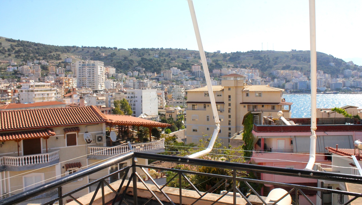 A panoramic view reveals the cityscape of Saranda, showcasing a blend of residential buildings and green hills. The blue waters of the sea glisten in the sunlight, with various structures framed by the balcony's railing in the foreground.
