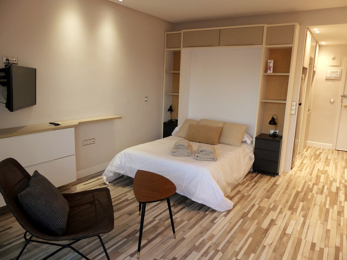 A well-lit room features a spacious bed with neatly arranged linens and two towels. A flat-screen television is mounted on the wall, while a small table and a chair provide additional seating. The light wood flooring contributes to the room’s airy feel.