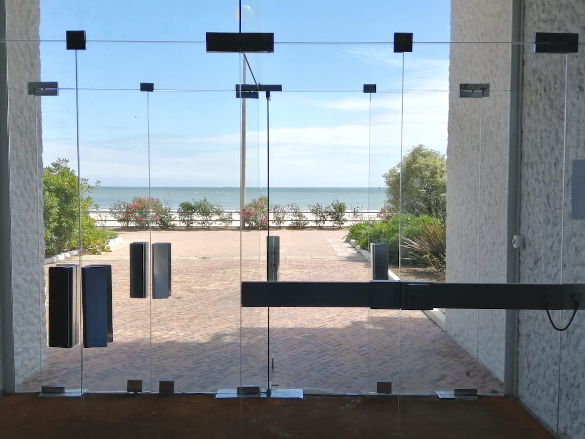 Transparent glass doors lead to a coastal view, with a path extending to the beach. The scene features greenery flanking the walkway and a clear blue sky overhead. The ocean is visible in the distance, creating a seamless indoor-outdoor transition.