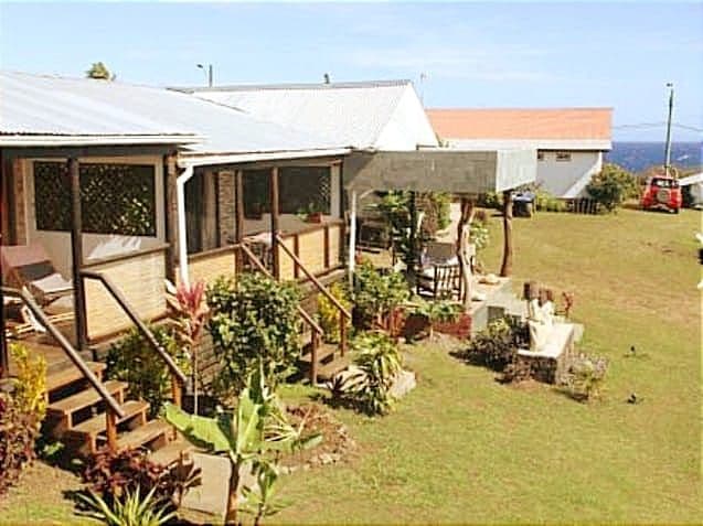 The exterior of the cabin features a welcoming porch with seating, surrounded by well-maintained gardens. A grassy area extends towards the horizon, where views of the ocean can be seen. A few vehicles are parked in the distance, contributing to the serene landscape.