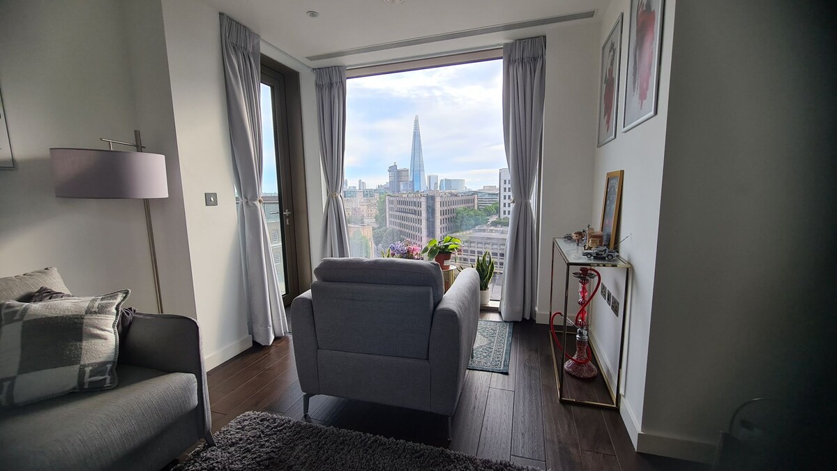 A comfortable living area features a view of London's skyline through large glass doors. The space is furnished with a light grey sofa and a small side table, while potted plants add a touch of greenery. Soft curtains frame the window overlooking the city.