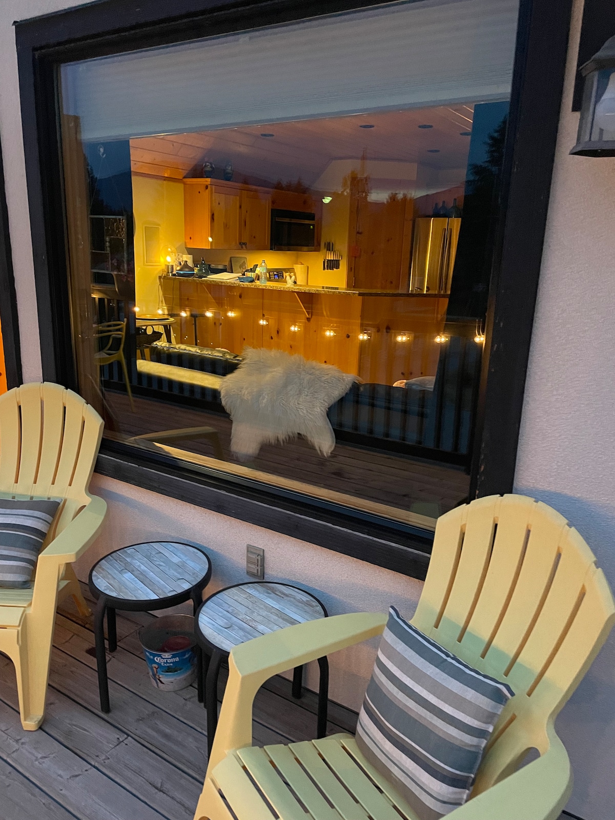 A cozy outdoor seating area includes two light yellow chairs and small round tables. A large window reflects the kitchen's warm glow, showcasing a modern wooden interior illuminated by soft lights. The balcony features a serene atmosphere suitable for relaxation.