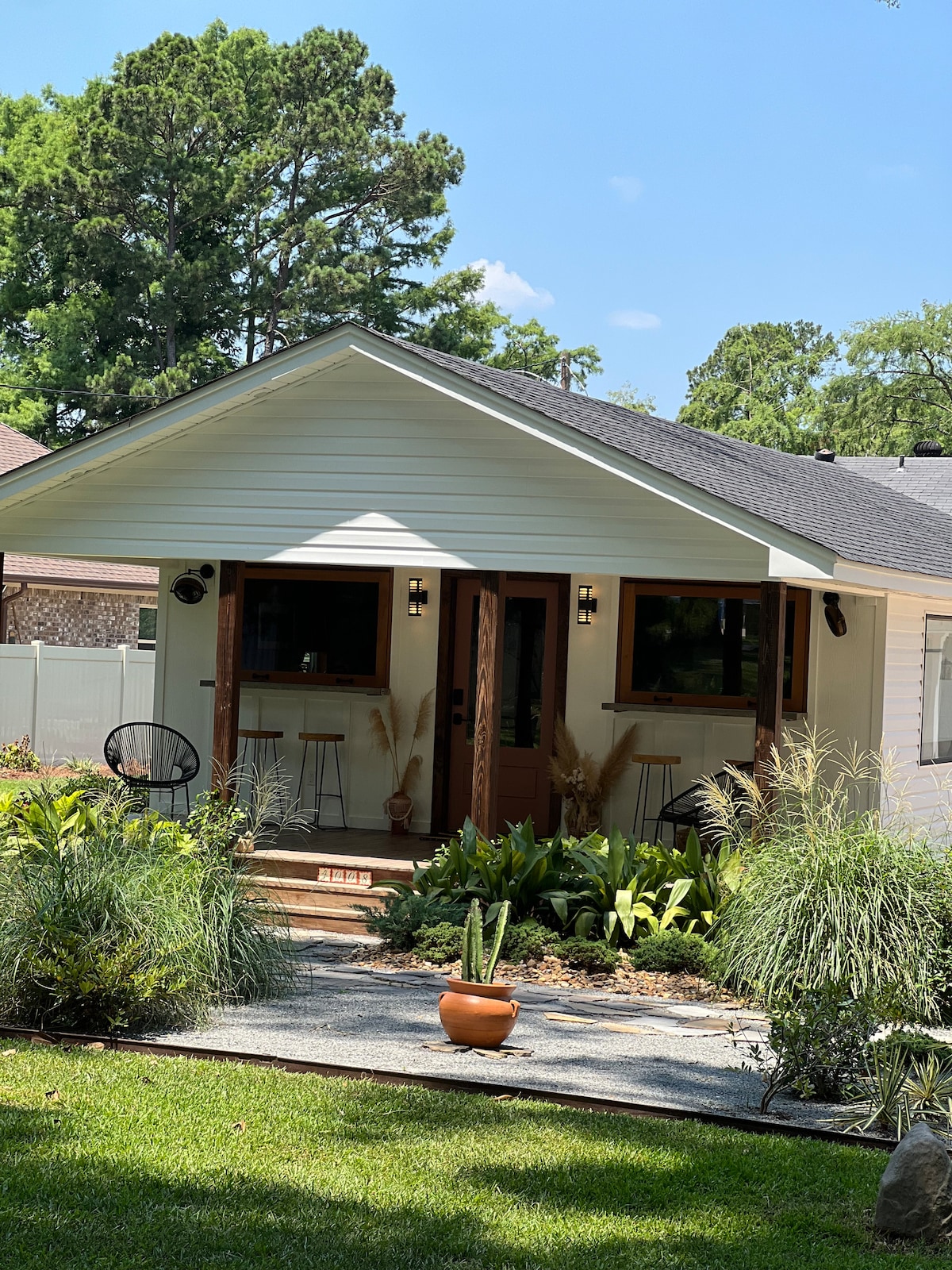 A cottage exterior is showcased, featuring a welcoming entrance with double doors. Lush greenery and arranged rocks lead to the home, while the porch area includes minimalist furniture. Bright sunlight highlights the clean lines of the structure against a backdrop of tall trees.