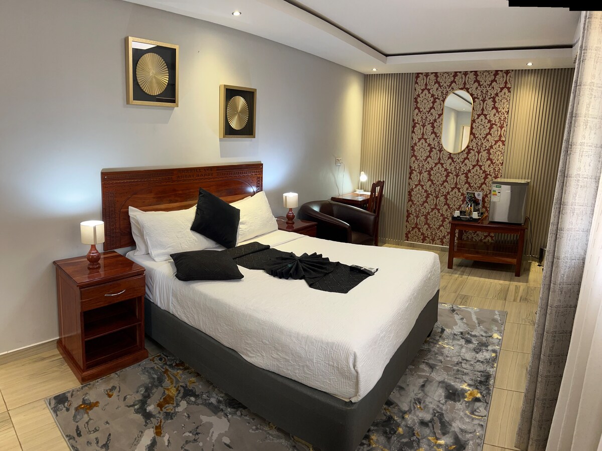 A well-appointed bedroom features a spacious bed with white linens and decorative black accents. Two bedside lamps provide soft illumination. A stylish armchair is positioned beside a small table, while a mini-fridge and a large mirror are visible against the wall adorned with patterned wallpaper.
