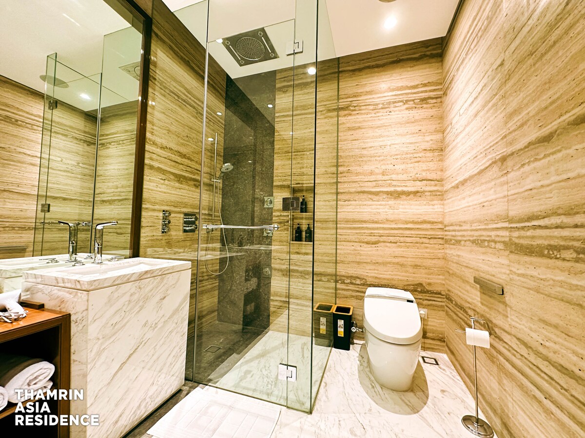 A modern bathroom features glass panels enclosing a spacious shower area and a sleek toilet. Marble countertops and warm textured walls create a sophisticated ambiance. Toiletries are neatly arranged on the shelves, with a towel rack and paper holder visible for convenience.