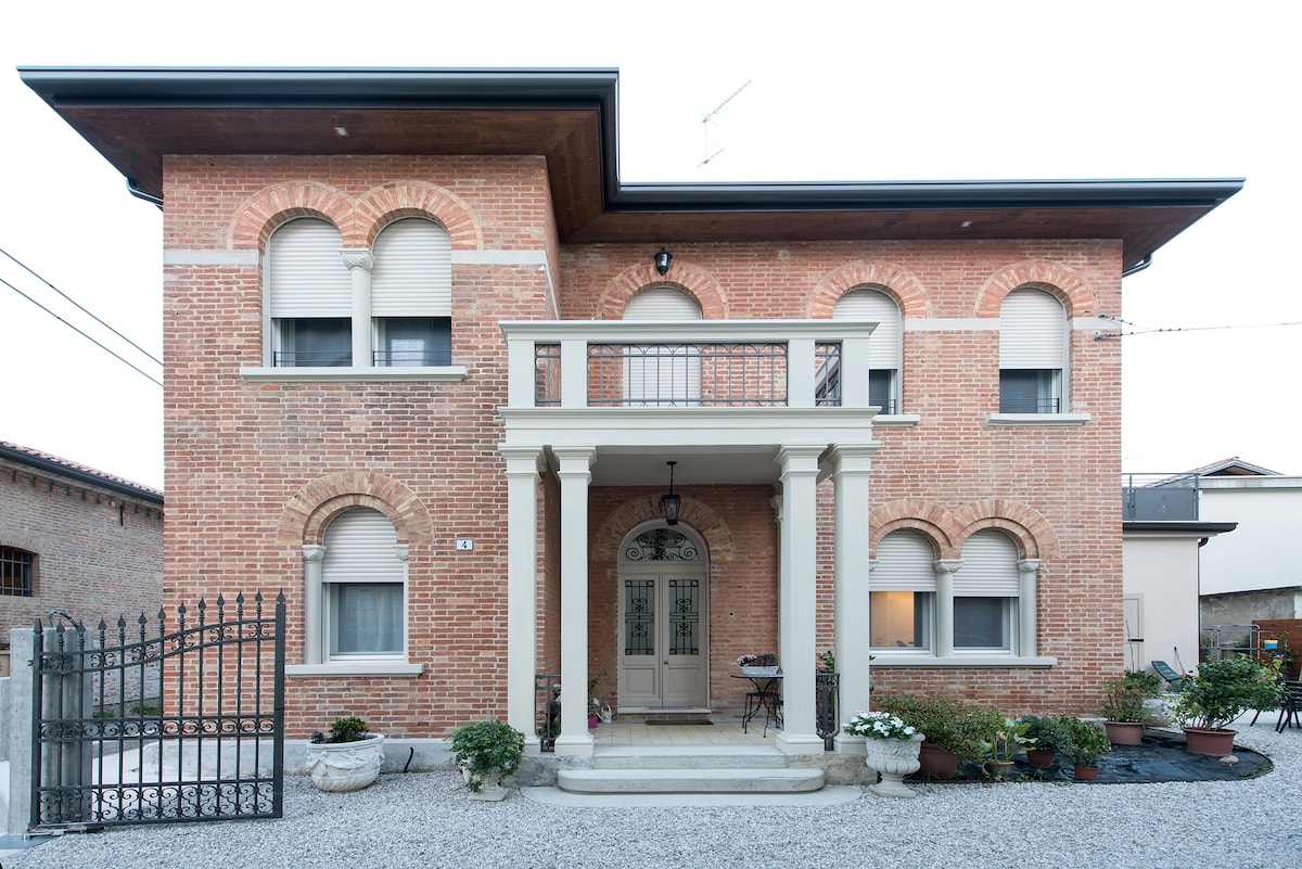 The exterior of a brick house is displayed, featuring arched windows and a prominent entrance. A covered porch supported by columns leads to the front door. Decorative plants line the entrance, and a gravel pathway adds texture to the environment.