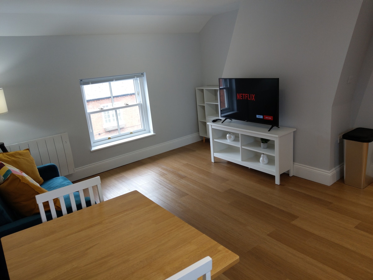 The living area features a comfortable blue sofa paired with a white dining table and chairs, creating a cozy space. A wall-mounted TV displays a Netflix logo, while natural light enters through the large window, enhancing the bright interior. Warm wood flooring complements the setting.