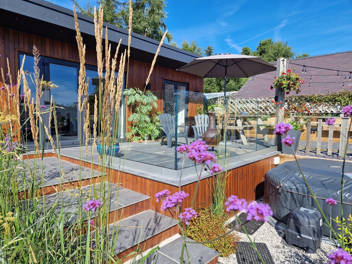 A private patio area is surrounded by lush greenery and flowers, leading to the cabin. Relaxing seating options include adirondack chairs and a large umbrella for shade. A hot tub is visible on the patio, enhancing the outdoor experience.