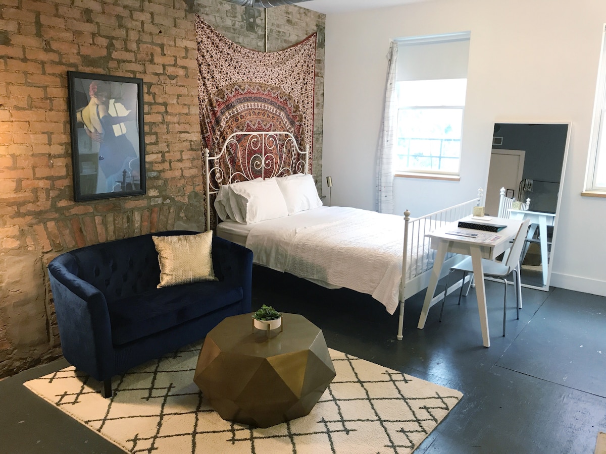 A studio apartment displays a blend of modern and vintage elements. A queen bed with crisp white bedding is positioned against a brick wall, complemented by a decorative tapestry above. A blue velvet couch sits near a geometric coffee table, while a small dining area is set by the window.