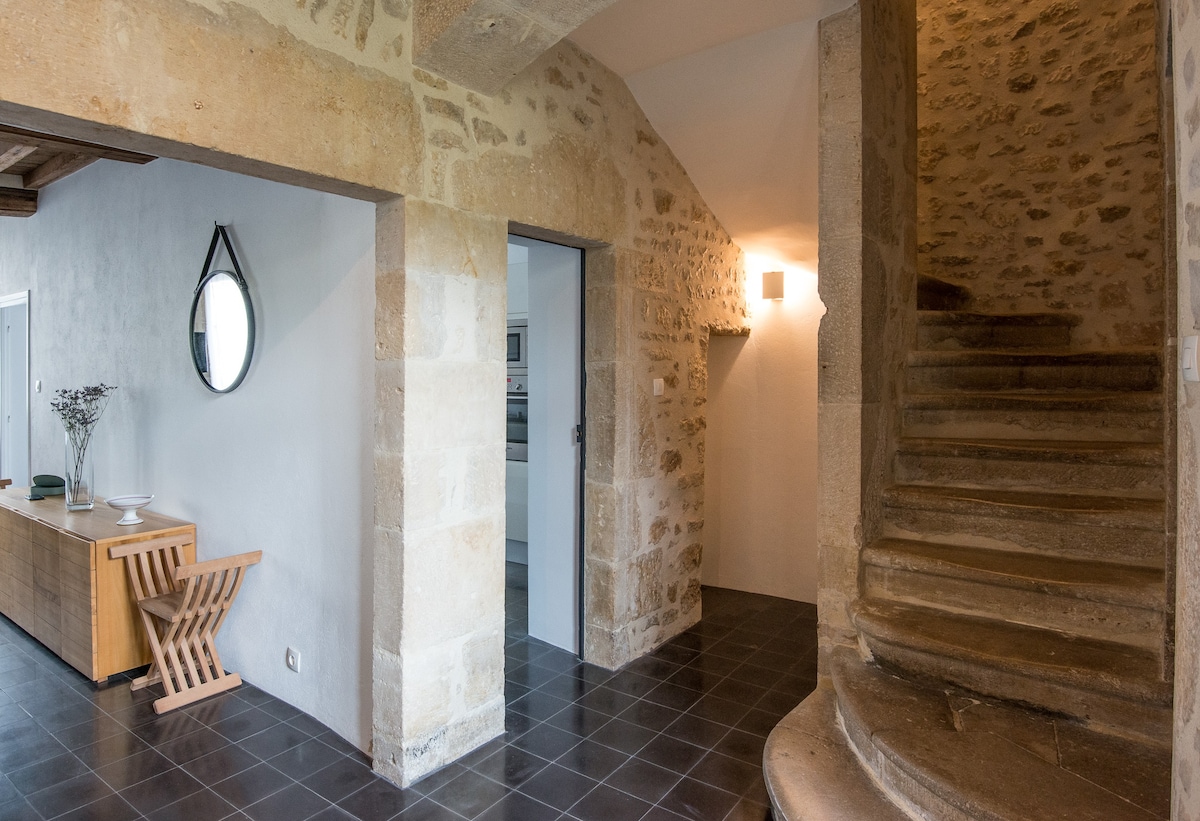 The entrance area features stone walls and a charming staircase crafted from natural stone. A wooden console table complements the space, alongside a circular mirror reflecting the light. Subtle lighting and a dark tile floor enhance the welcoming ambiance.