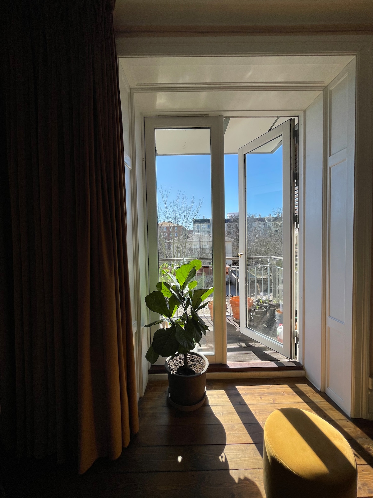 A set of double doors opens to a sunny balcony, framed by warm curtains. A potted plant sits at the base, placed on the polished wooden floor. Clear blue skies and buildings can be seen through the doorway, enhancing the inviting atmosphere.