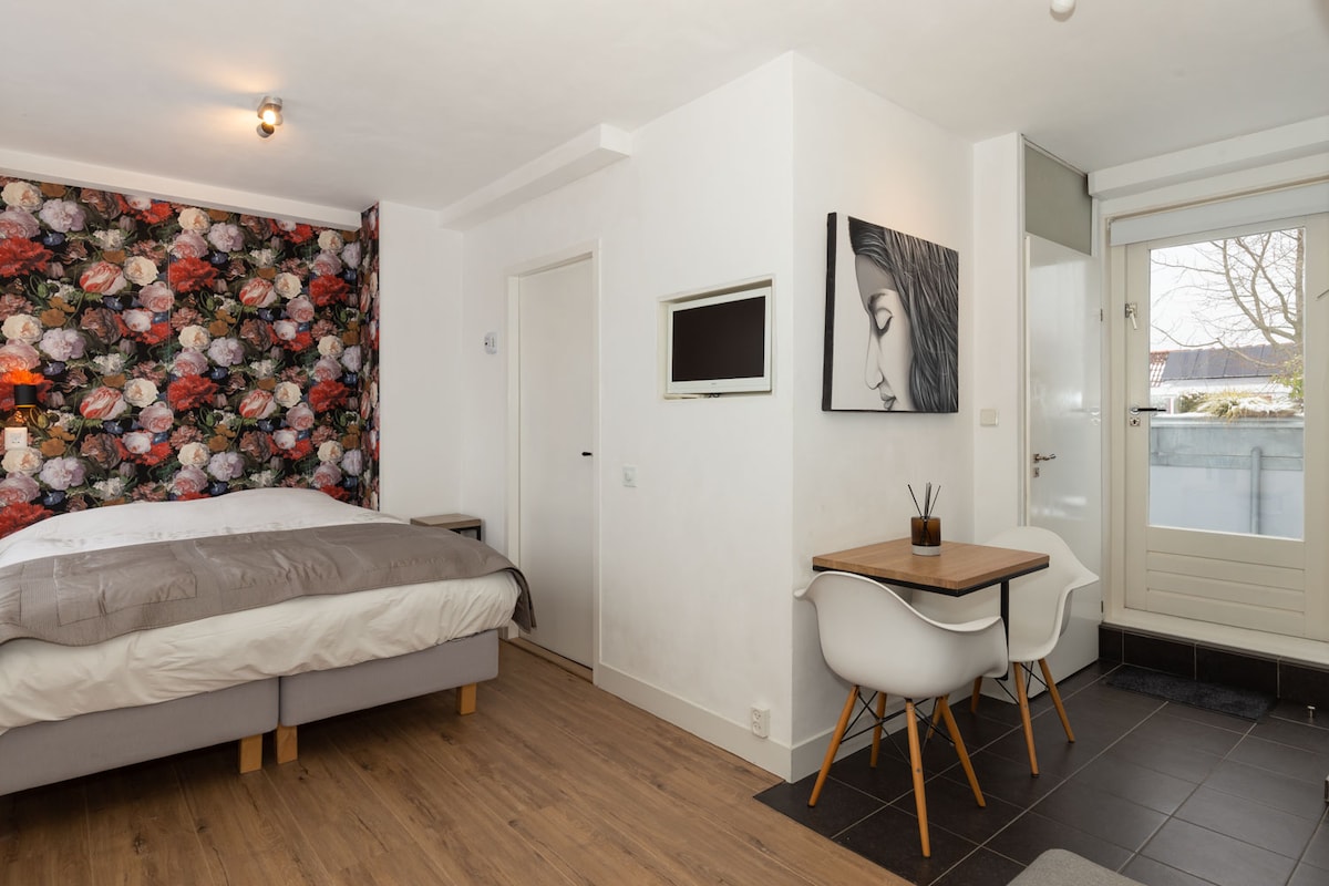 A bright studio features a spacious bed with light bedding against a floral accent wall. A minimalist dining area is situated beside a large window, offering natural light. A mounted screen and a door leading to an outdoor space are also visible, enhancing the functionality of the space.