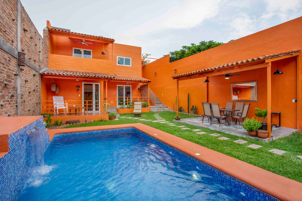 A vibrant orange house is depicted with a swimming pool in the foreground. The pool is surrounded by lush green grass and bordered by small stone pathways. Several lounge chairs and a dining area with a table and chairs are visible, providing space for outdoor relaxation.