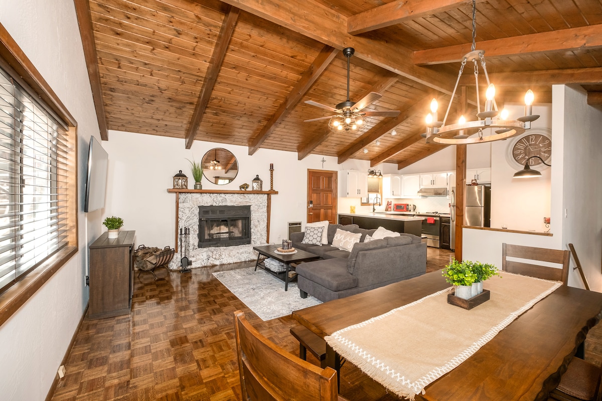 A spacious living area is highlighted by vaulted wooden ceilings and rich parquet flooring. A stone-surrounded fireplace serves as a focal point, complemented by a sectional sofa and a wooden dining table. Natural light filters through a large window, creating a welcoming ambiance.