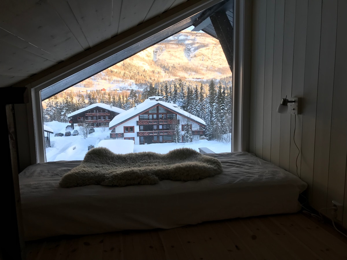 A cozy sleeping area features a large mattress positioned by a slanted window. Snow-covered mountains and a cluster of traditional wooden buildings are visible outside, surrounded by tall evergreen trees. A soft, woven throw blanket is casually placed over the mattress.