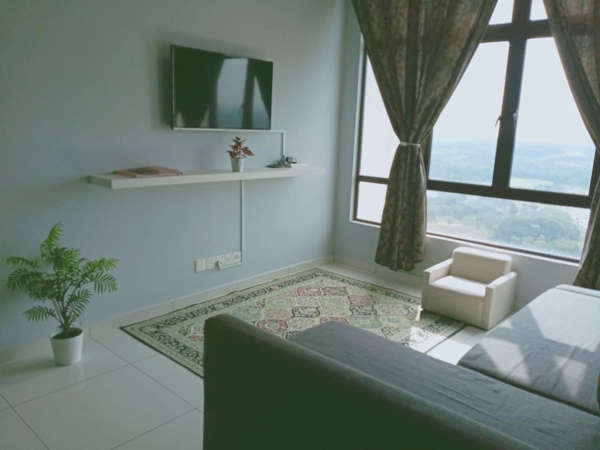 A modern living area features a small seating arrangement with a light-colored chair and a plush sofa. A wall-mounted television is positioned above a sleek shelf, while large windows draped in sheer curtains offer a view of the landscape outside.