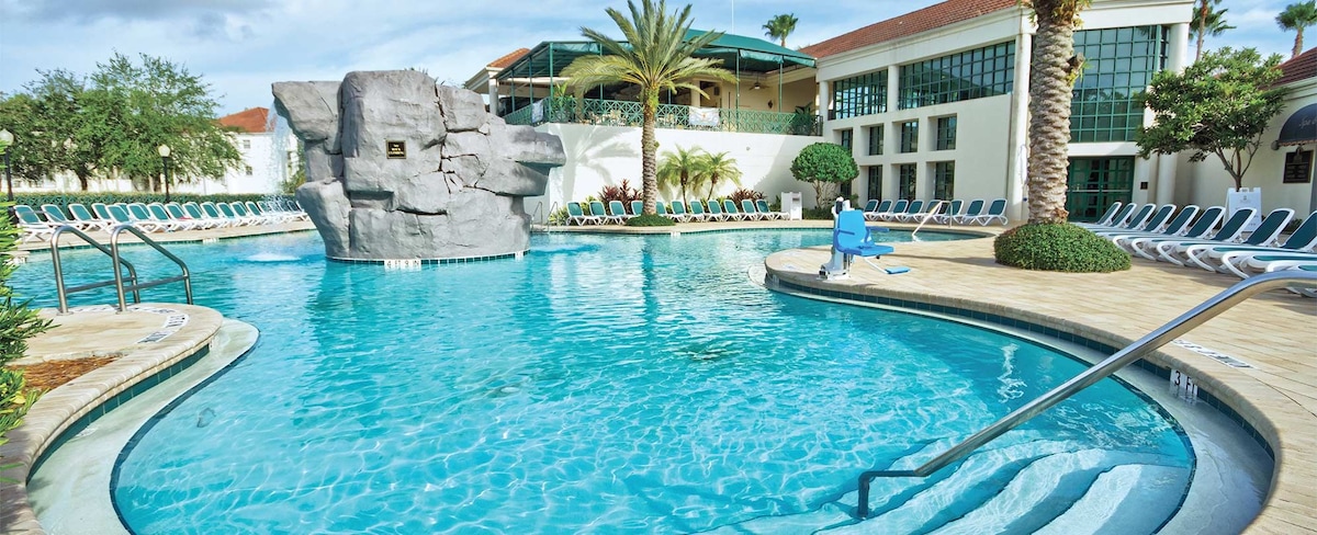 A tranquil pool area is surrounded by lush greenery and palm trees. The water appears clear and inviting, with a rock formation creating a natural aesthetic. Lounge chairs are positioned around the pool, providing ample space for relaxation.