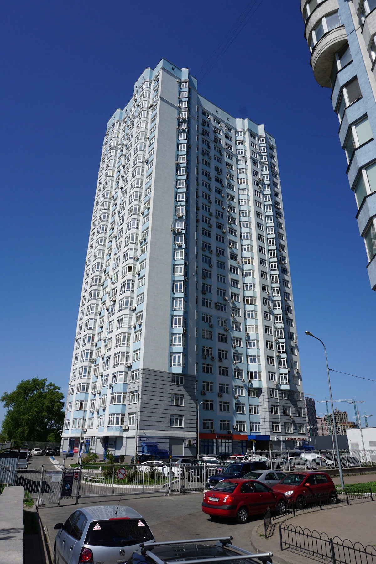 A tall modern building is shown against a clear blue sky. The exterior features a mix of pastel colors and numerous windows, allowing natural light to illuminate the interiors. Several cars are parked in the foreground, with surrounding greenery adding a touch of nature.