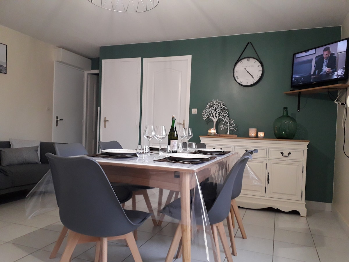 A dining area is presented with a large wooden table surrounded by six grey chairs. Clear glasses and elegant tableware are neatly arranged. A television is mounted on the wall, and a green accent wall adds a touch of color. A clock and decorative pieces are visible.