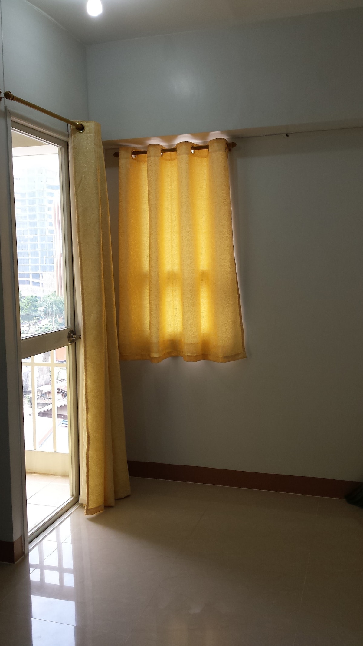 A bright corner of the room features a glass door leading to a small balcony, surrounded by light-colored walls. Soft yellow curtains frame the window, allowing gentle daylight to filter through. The tiled floor reflects the warm light, enhancing the room's spacious feel.