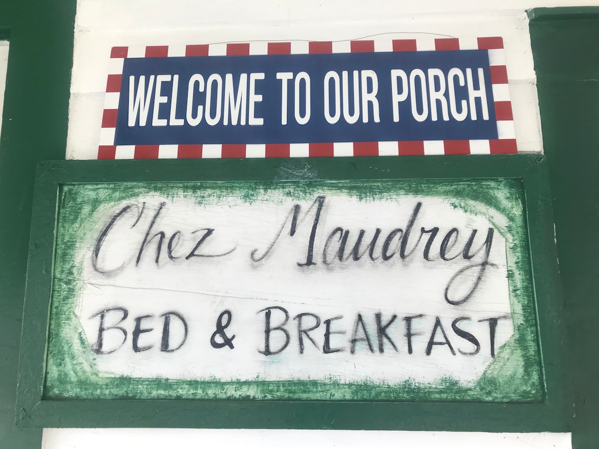 A welcoming porch area is highlighted by a decorative sign reading ‘WELCOME TO OUR PORCH.’ Below, a wooden sign displays ‘Chez Maudrey Bed & Breakfast’ in an elegant script, framed by green accents that complement the inviting entrance.