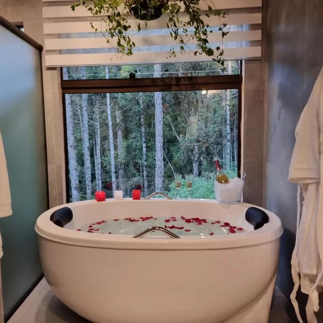 A spacious bathtub is featured, surrounded by candles and adorned with rose petals. A large window overlooks a serene forest, allowing natural light to brighten the space. Soft bathrobes hang nearby, enhancing the relaxing atmosphere.