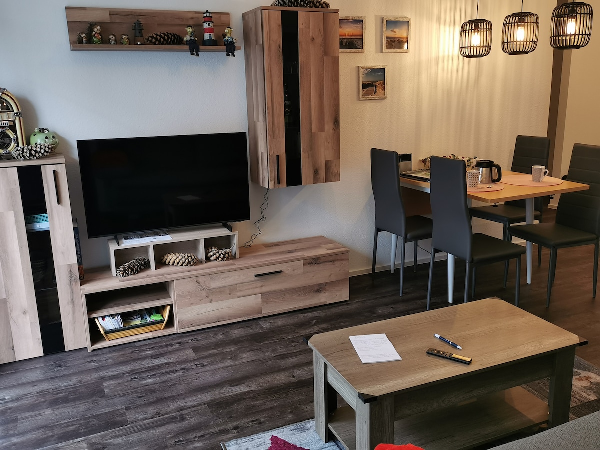 A modern living area features a flat-screen television mounted on a wooden unit, complemented by a matching coffee table. A dining space is visible, equipped with a wooden table surrounded by four grey chairs. Decorative items add a subtle touch to the overall design.