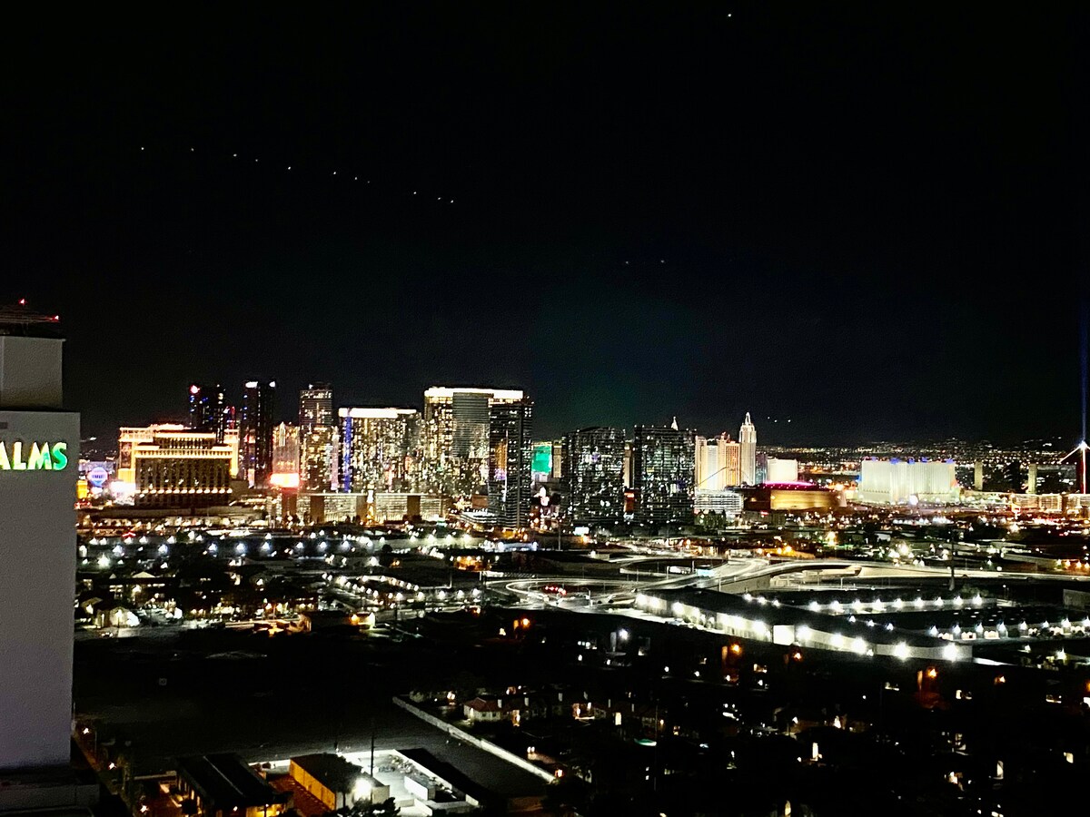 A night view of the Las Vegas Strip is displayed, showcasing illuminated skyscrapers and vibrant signage. The contrast between the bright city lights and the dark sky creates a striking visual. The Palms Hotel sign sits in the foreground, reflecting the lively atmosphere of the area.