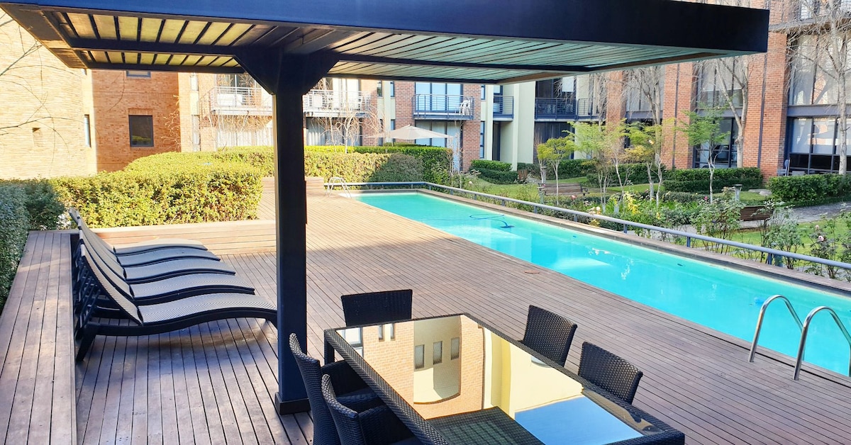 An outdoor area features a sleek dining table surrounded by stylish chairs, shaded by a pergola. A long, rectangular swimming pool is visible alongside sun loungers arranged on a wooden deck. Lush greenery provides a natural backdrop to the serene setting.