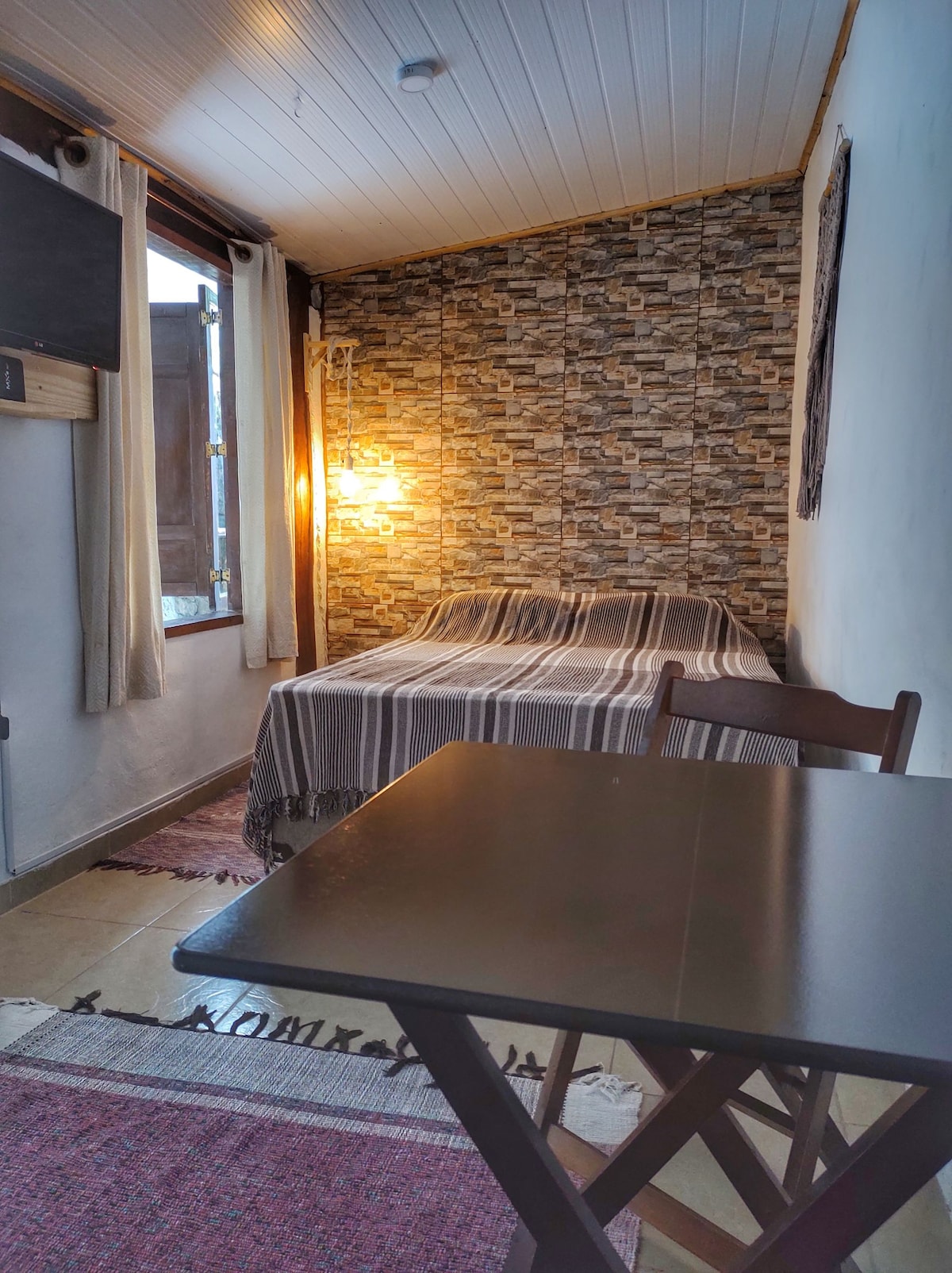 A cozy sleeping area is featured, with a striped blanket on a spacious bed against a patterned accent wall. Natural light filters through the window, complemented by warm lighting from a nearby lamp. A small table with folding chairs is also present.