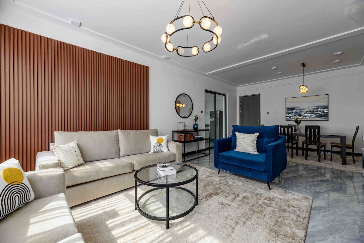 The living area features a combination of a light-colored sofa and a blue armchair, complementing a glass coffee table on a textured rug. Wooden paneling adds warmth, while a round chandelier and wall art enhance the ambiance. A dining area with a dark table is visible in the background.
