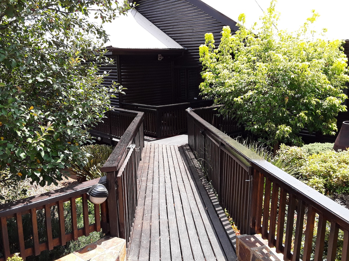 A wooden pathway leads through lush greenery, connecting a scenic log cabin with surrounding gardens. The pathway is flanked by various plants and a few trees, creating a natural border that enhances the tranquil setting.