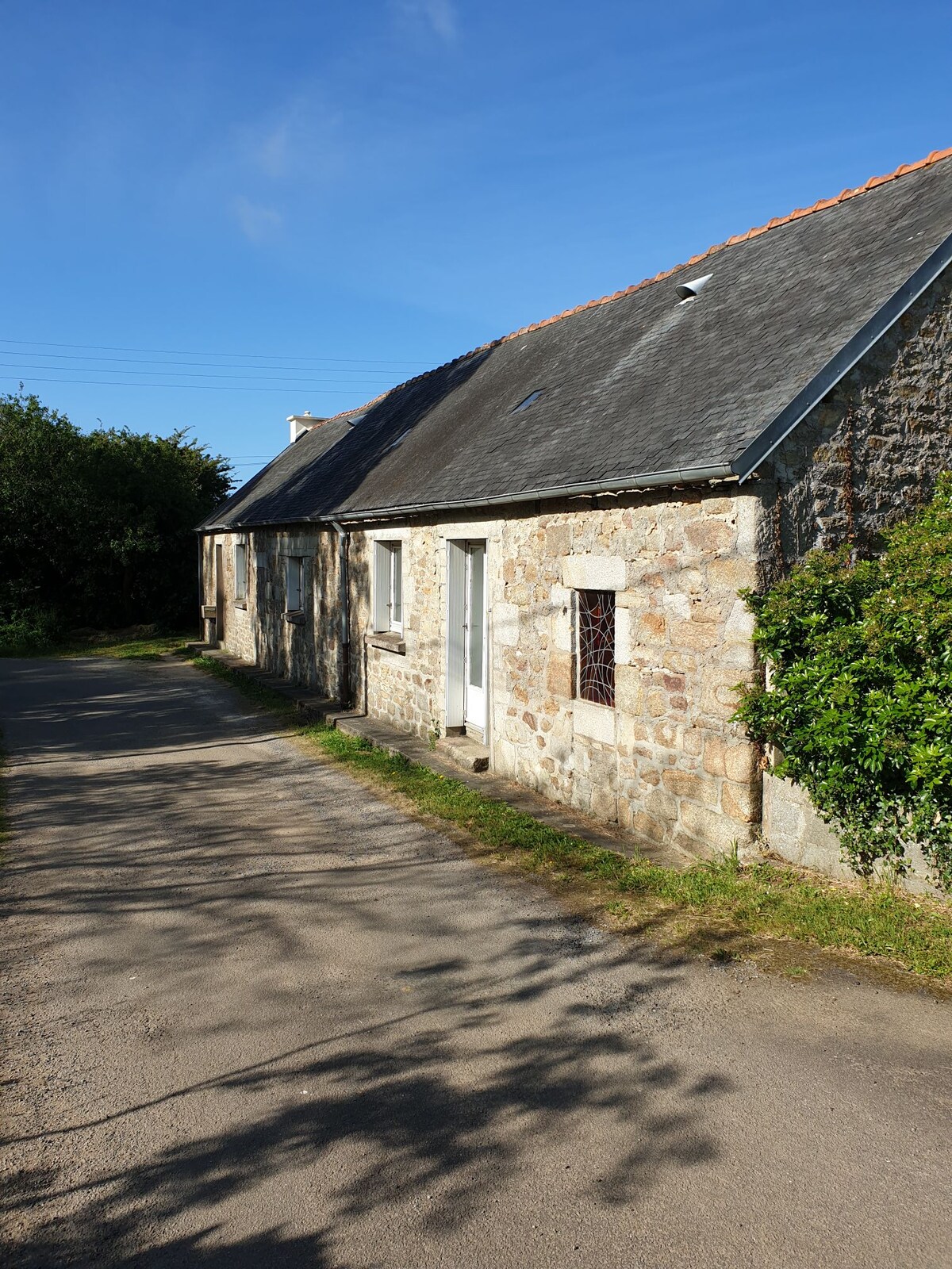 A neo-Breton house is pictured along a tranquil pathway. The stone exterior features a mix of light and dark tones, with a slate roof and white door. Green foliage on either side adds to the rural charm, complemented by a clear blue sky above.