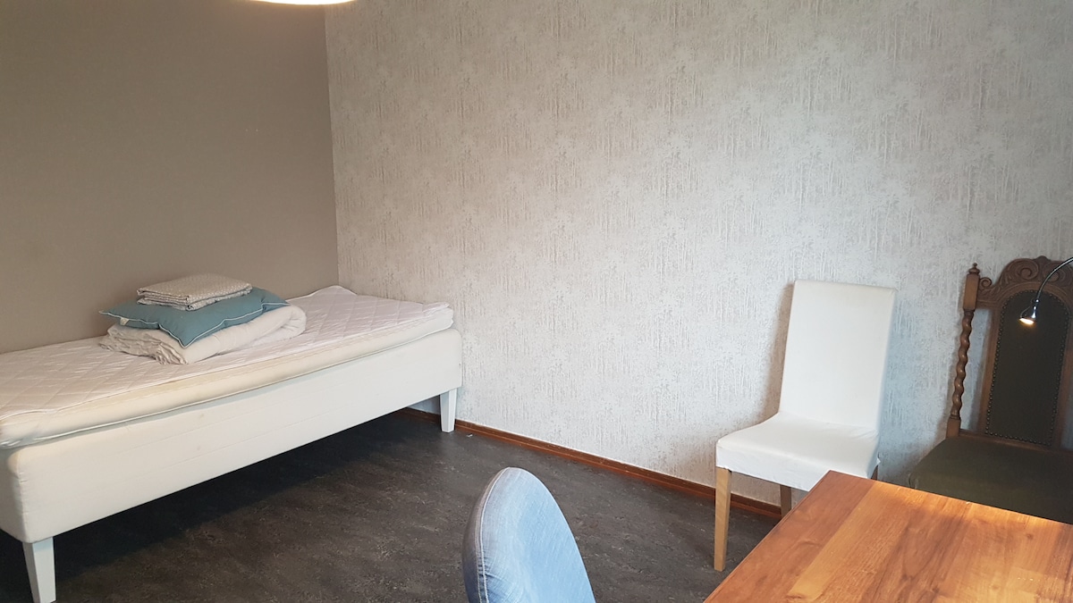 A furnished room features a single bed with folded linens and a soft blue blanket. A white chair and a wooden table are visible, adding to the room's functionality. The walls are decorated with a light, textured wallpaper, and the floor is finished with dark tiles.