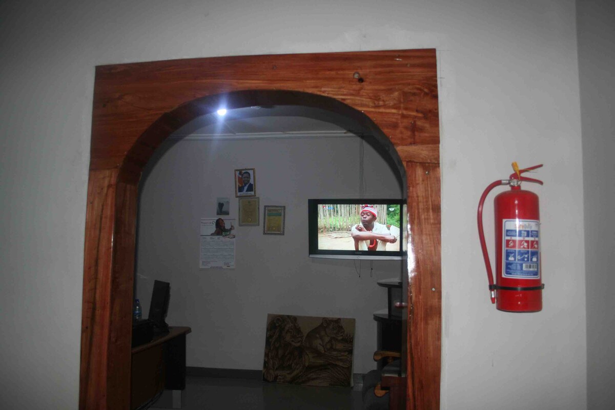 An arched wooden doorway leads into a well-lit room featuring a television mounted on the wall. A fire extinguisher is prominently displayed next to the entrance for safety. Notable decorations include framed images and documents on the walls.